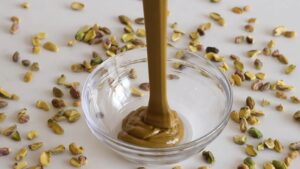 A Smooth, Rich Pistachio Paste Being Poured Into a Glass Bowl, Surrounded by Scattered Pistachios