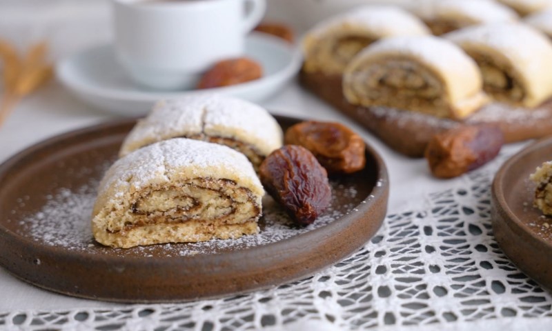 Powdered sugar-dusted pastry rolls on a ceramic plate, accompanied by dates