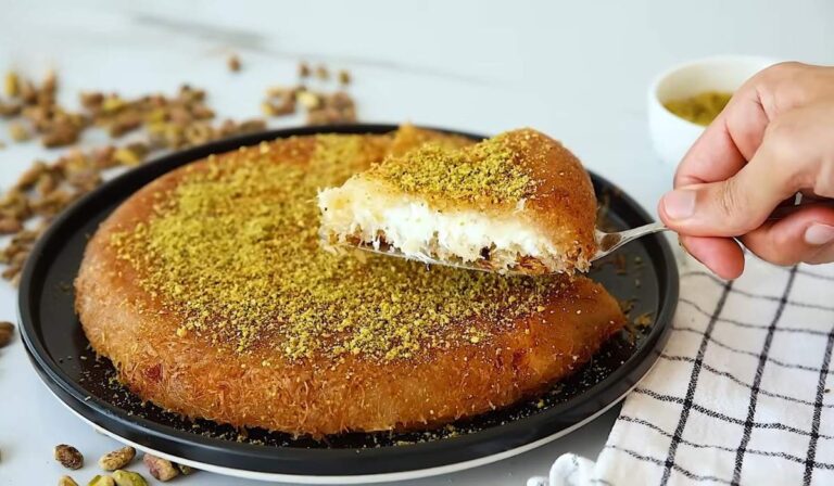 Slice of kanafa being lifted from a round tray topped with crushed pistachios