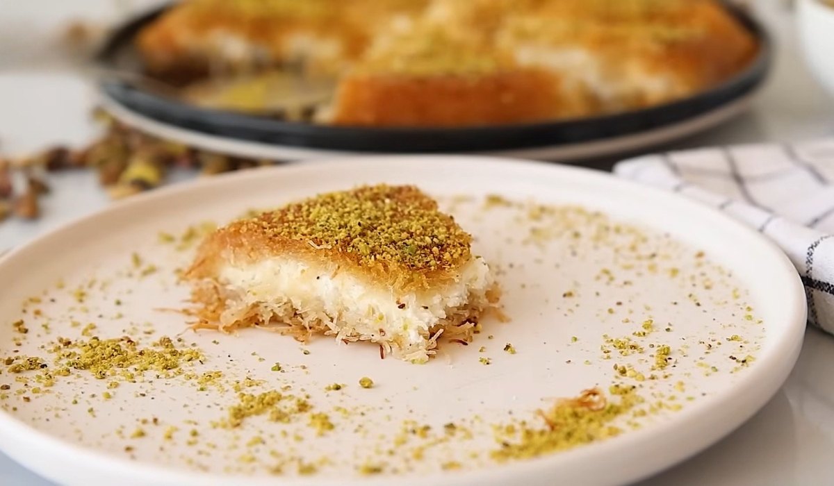 Single piece of kanafa on a plate with pistachio crumbs and tray in the background
