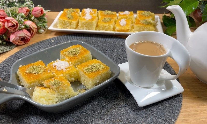 A cozy setup featuring a gray plate of yellow cake squares garnished with nuts, a white cup of tea, a teapot, and pink flowers on a gray mat