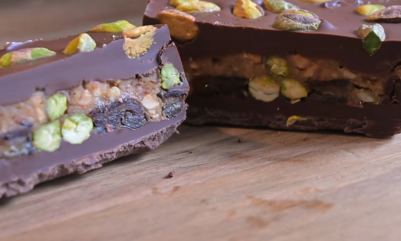 Close-up of a chocolate bar cut in half, revealing layers of nuts and dried fruits like pistachios and figs, on a wooden surface