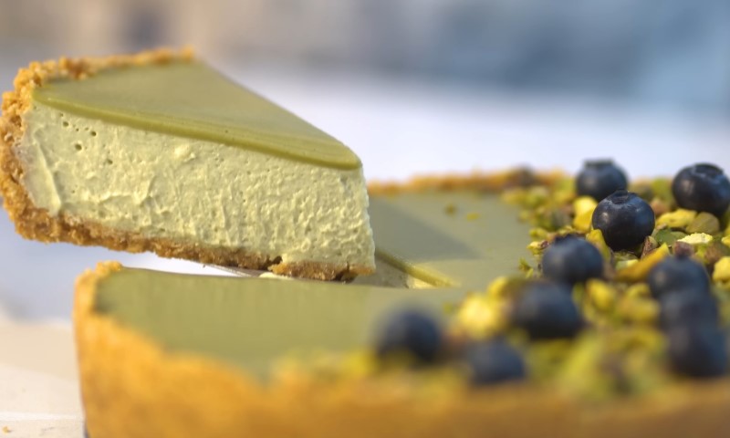 A creamy pistachio cheesecake slice being lifted from a whole cake
