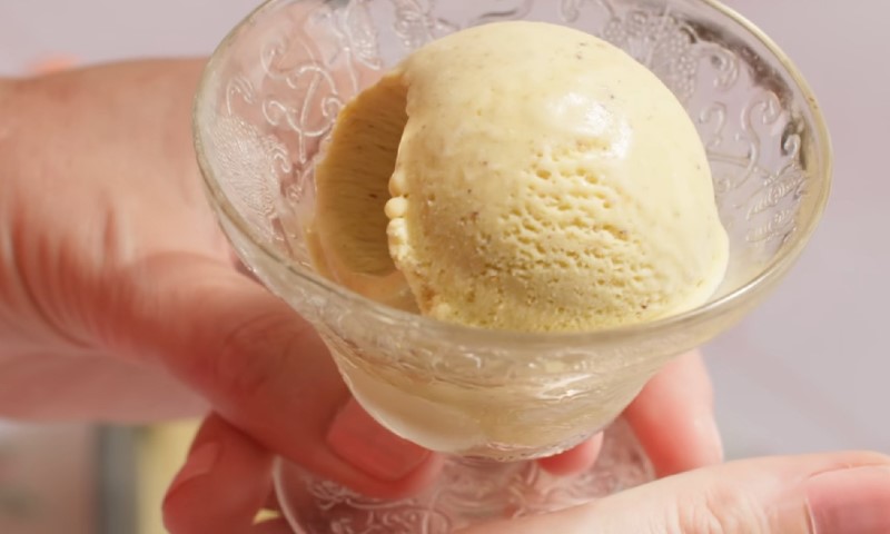 A hand holds a glass dish with a scoop of creamy, beige ice cream