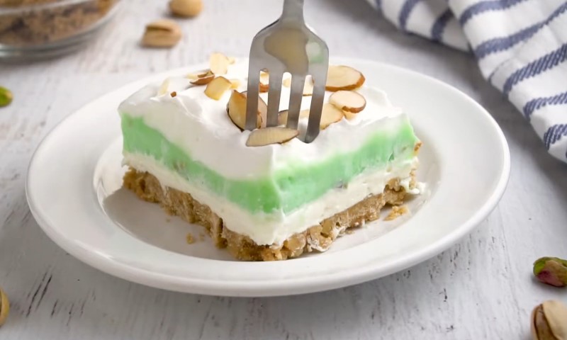 A creamy dessert slice with green and white layers topped with almond slices on a white plate. A fork is poised above, set on a rustic table