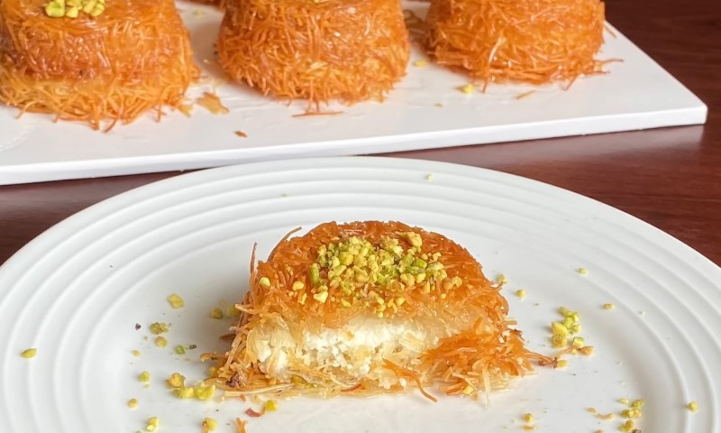 A close-up of a plate with a single piece of golden-brown knafeh topped with crushed pistachios, showing a cheese filling